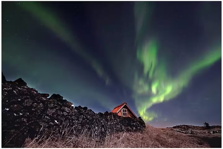 Aurora borealis northern lights swirling over a small red cabin and stone wall in Iceland at night