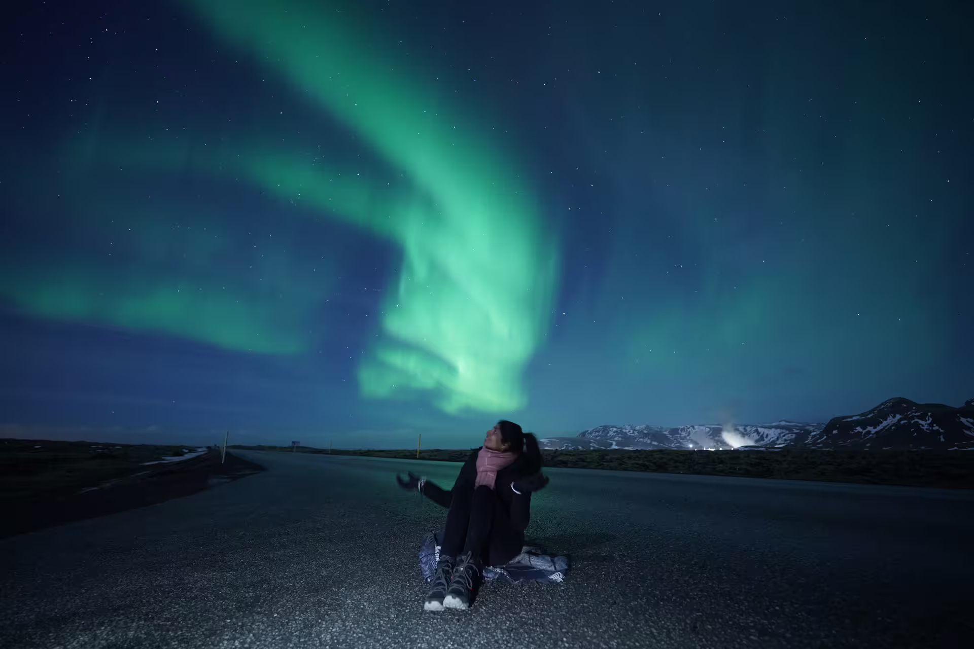 Woman sitting on road gazing at vibrant green aurora borealis over snowy Icelandic mountains at night