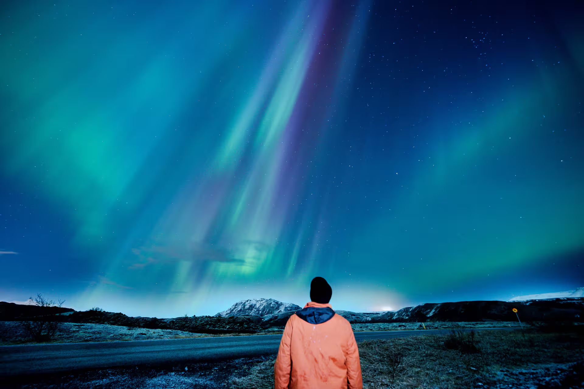 Person in orange jacket watching vibrant green and purple northern lights over Icelandic mountains at night