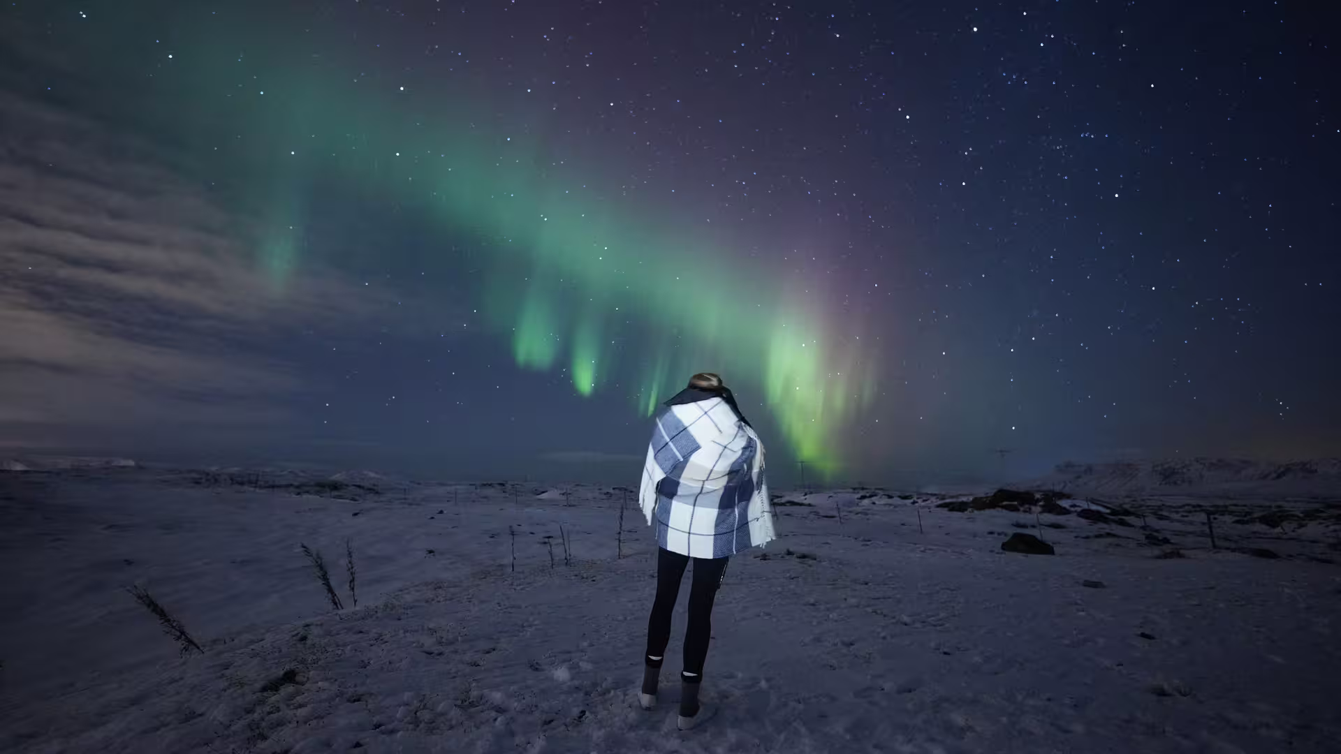 Person wrapped in plaid blanket watching vibrant green northern lights over snowy Iceland landscape at night