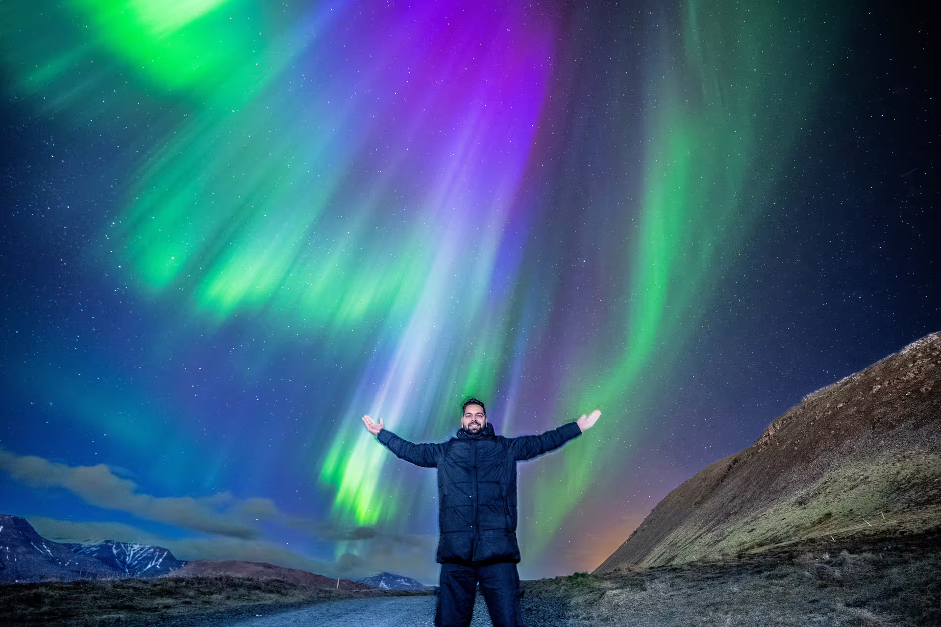 Man with arms outstretched under vivid green and purple northern lights in Iceland's night sky