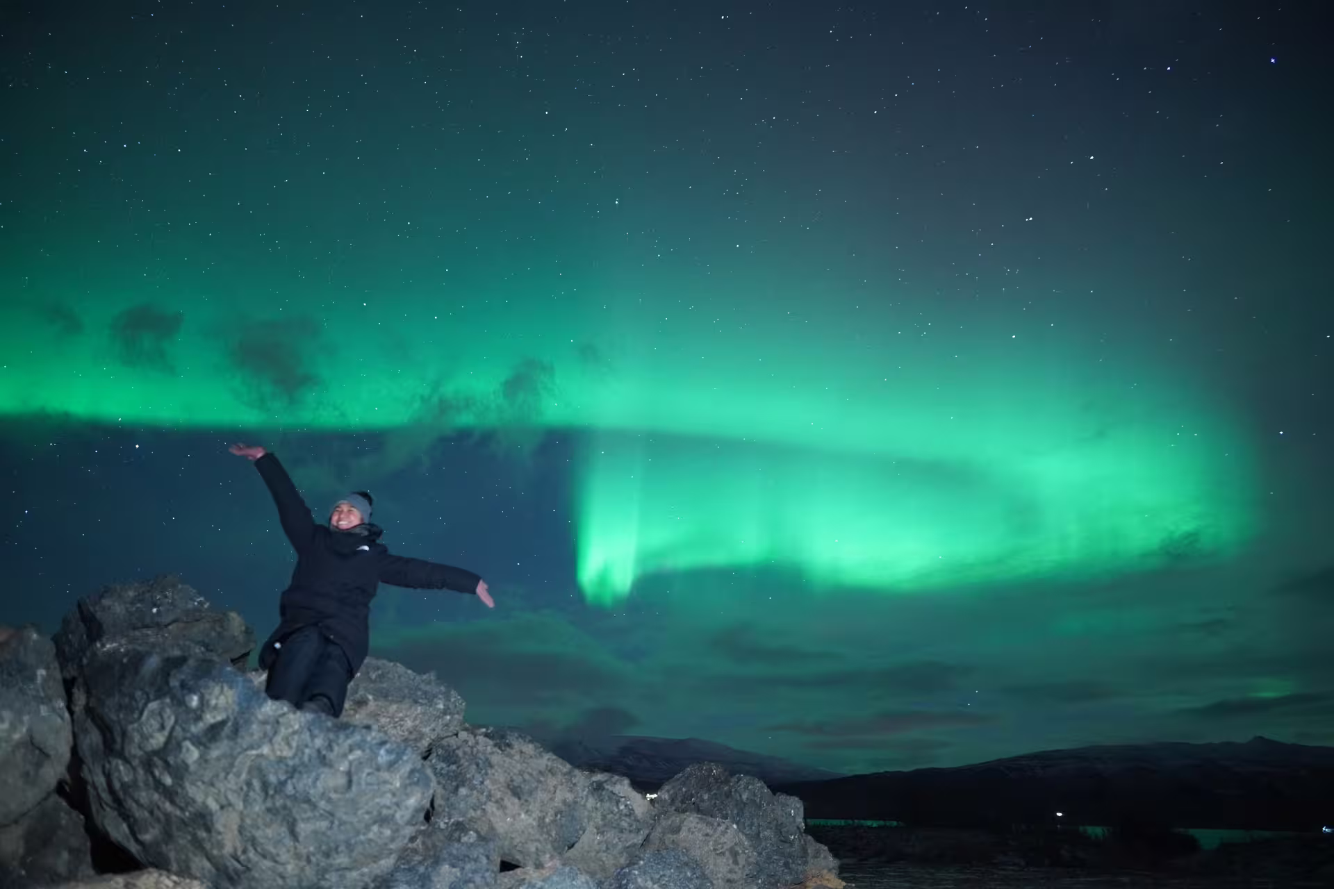 Person sitting on rocks with arms outstretched beneath vivid green northern lights in Iceland night sky