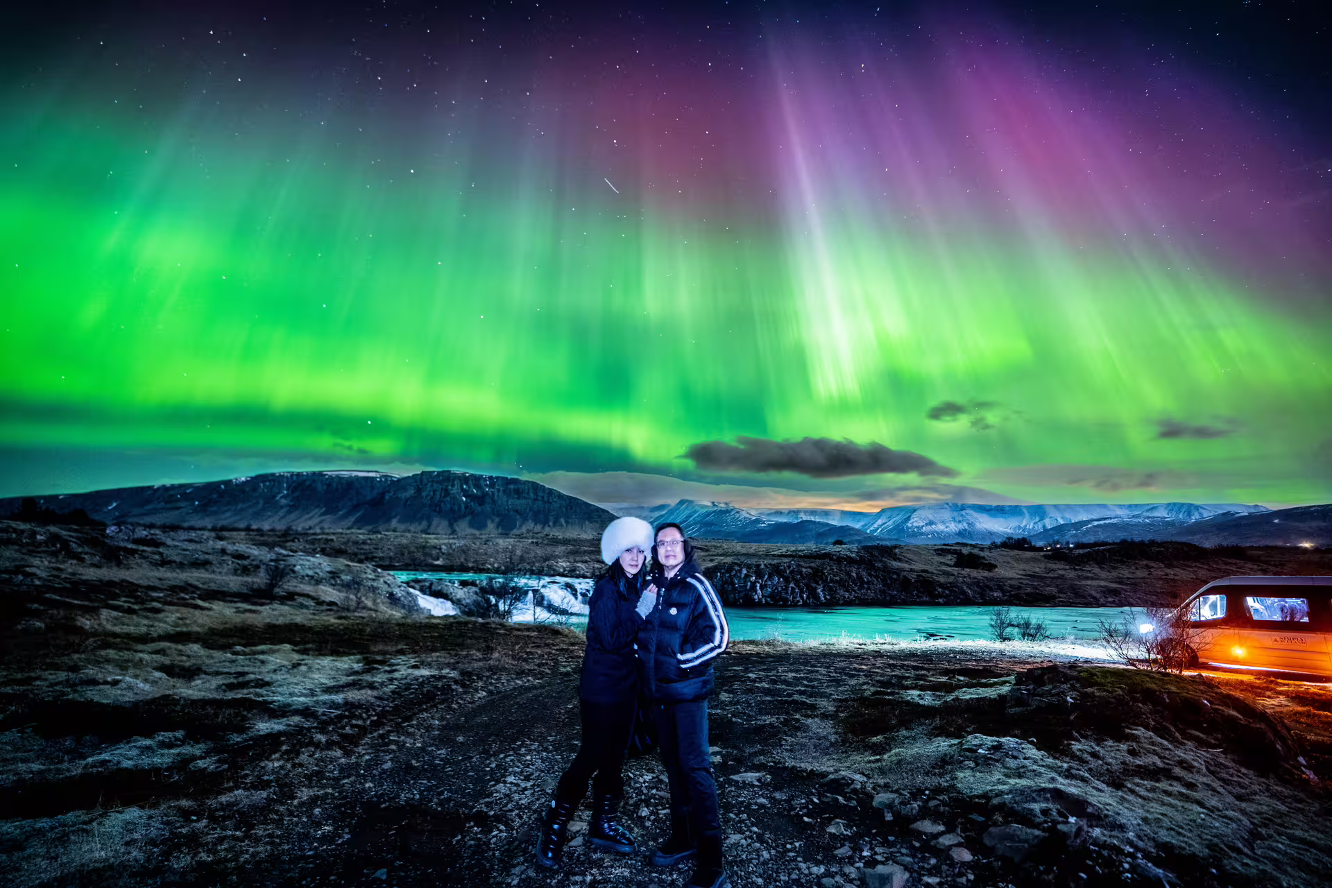 Two travelers posing under vibrant green and pink northern lights over Icelandic mountains at night