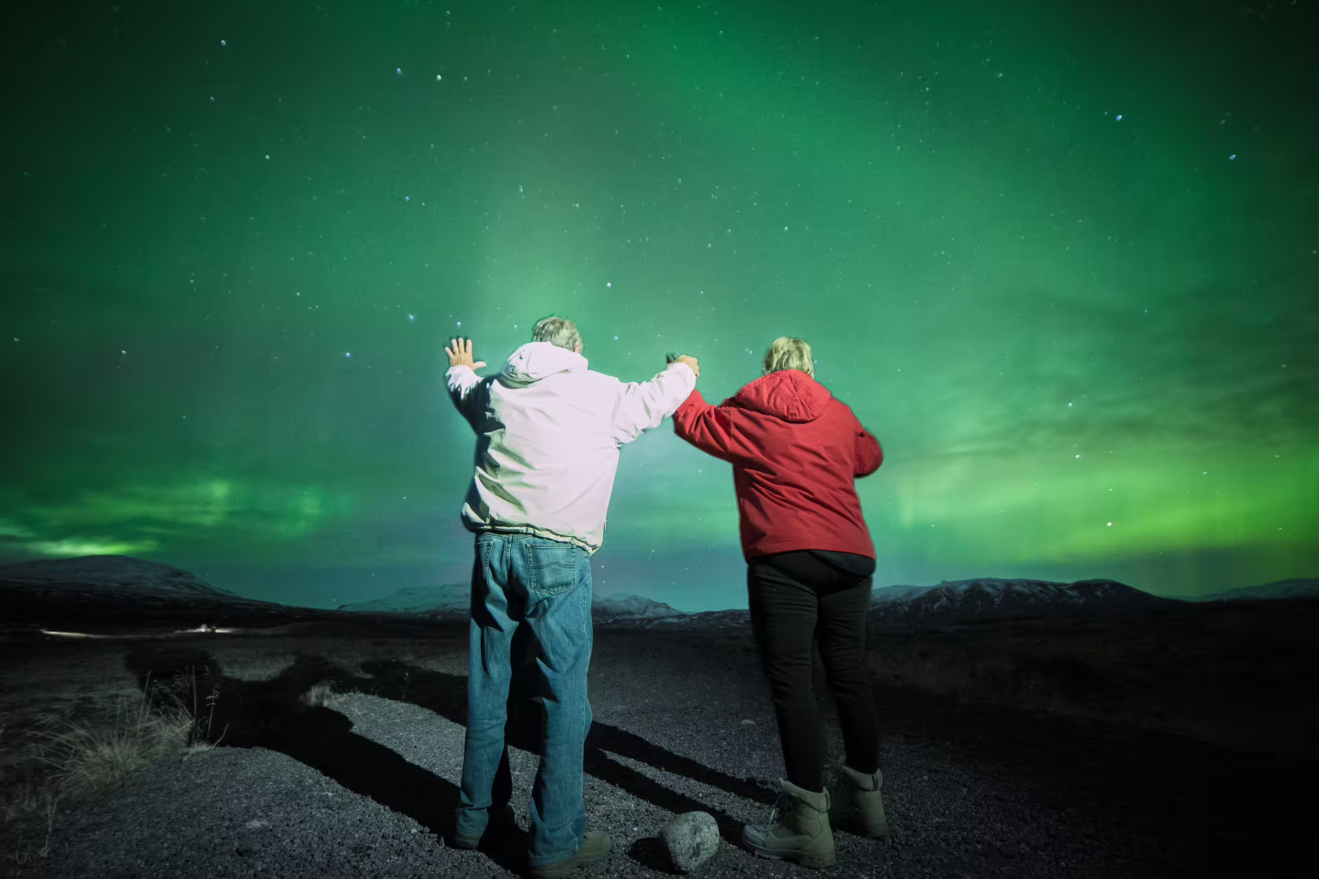 Two people holding hands watching vibrant green northern lights over Iceland's dark mountainous landscape at night