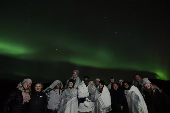 Group of tourists wrapped in blankets smiling under vibrant green northern lights in Iceland night sky