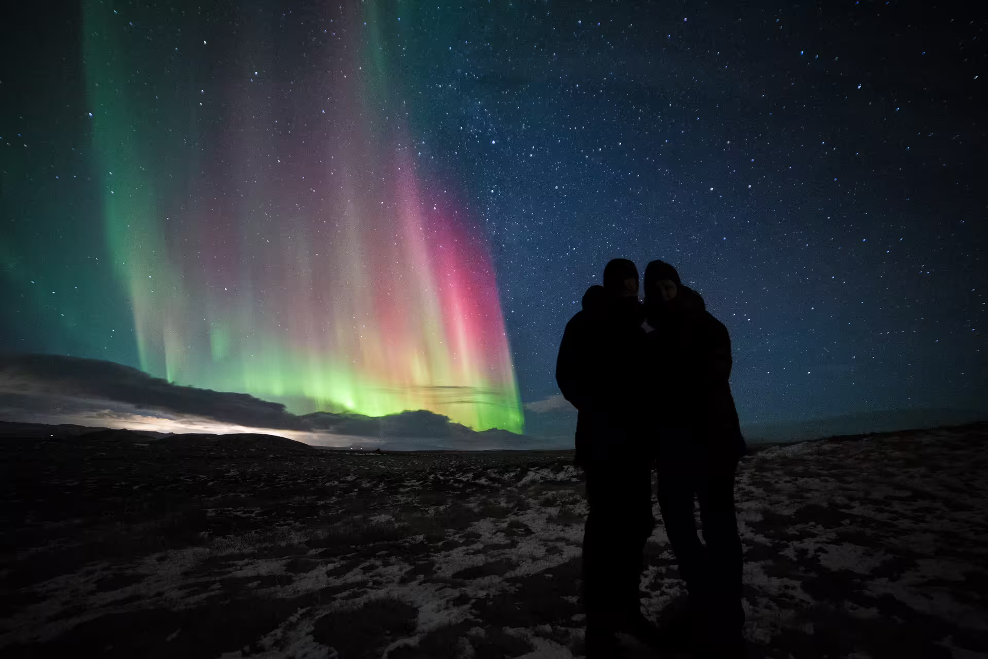 Two people silhouetted against vibrant green and pink northern lights over snowy Iceland landscape at night