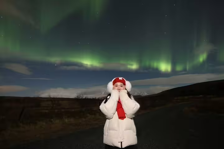 Woman in white puffer jacket and red hat posing under vibrant green northern lights in Iceland