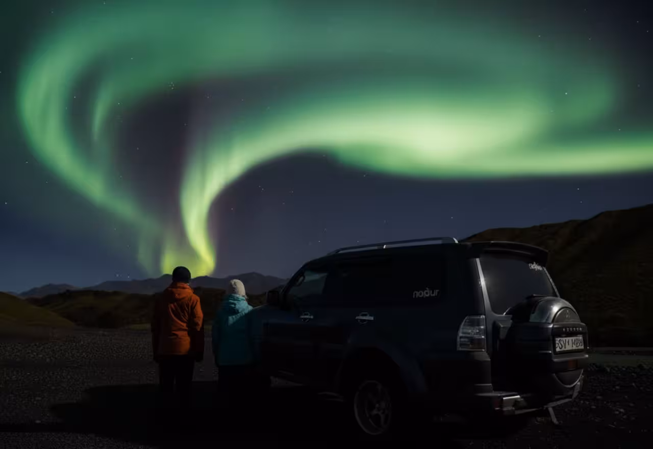 Two people watching vibrant green northern lights swirling over Iceland mountains beside a Super Jeep at night