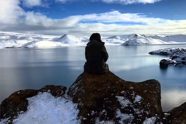 Person sitting on snowy rock overlooking calm blue lake with snow-capped mountains in Iceland