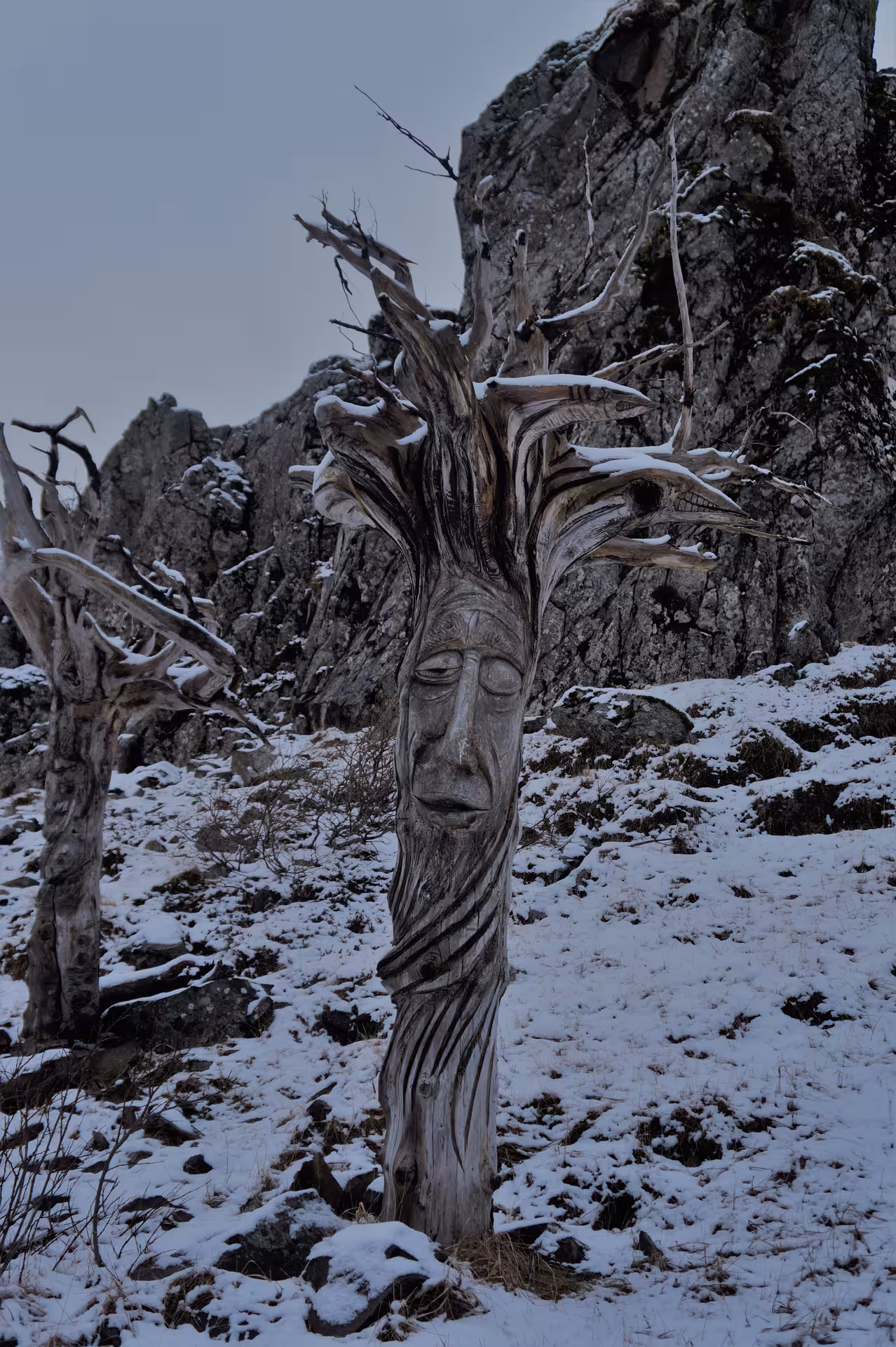 Carved wooden face sculpture on a dead tree trunk in a snowy Icelandic mountain landscape