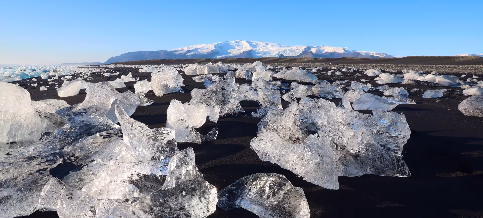 Crystal ice chunks scattered on black sand beach with snow-capped Icelandic glacier in background
