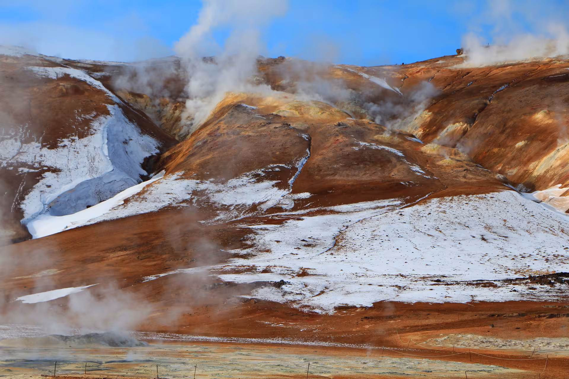 Steaming geothermal hills with rust-red rhyolite mountains and snow patches on Iceland Ring Road tour
