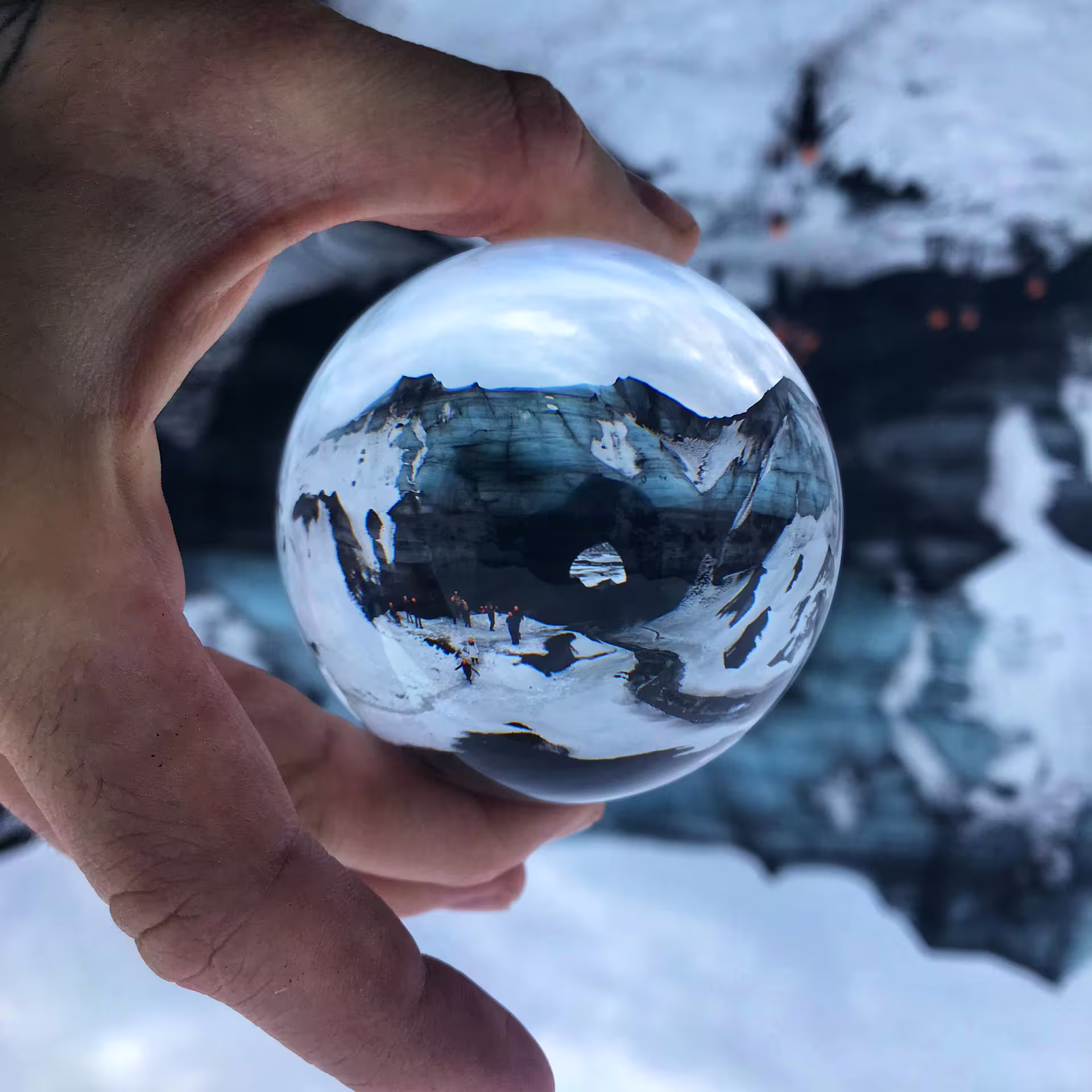 Crystal ball held by hand reflecting snowy Iceland mountains and tourists on glacier during ring road tour