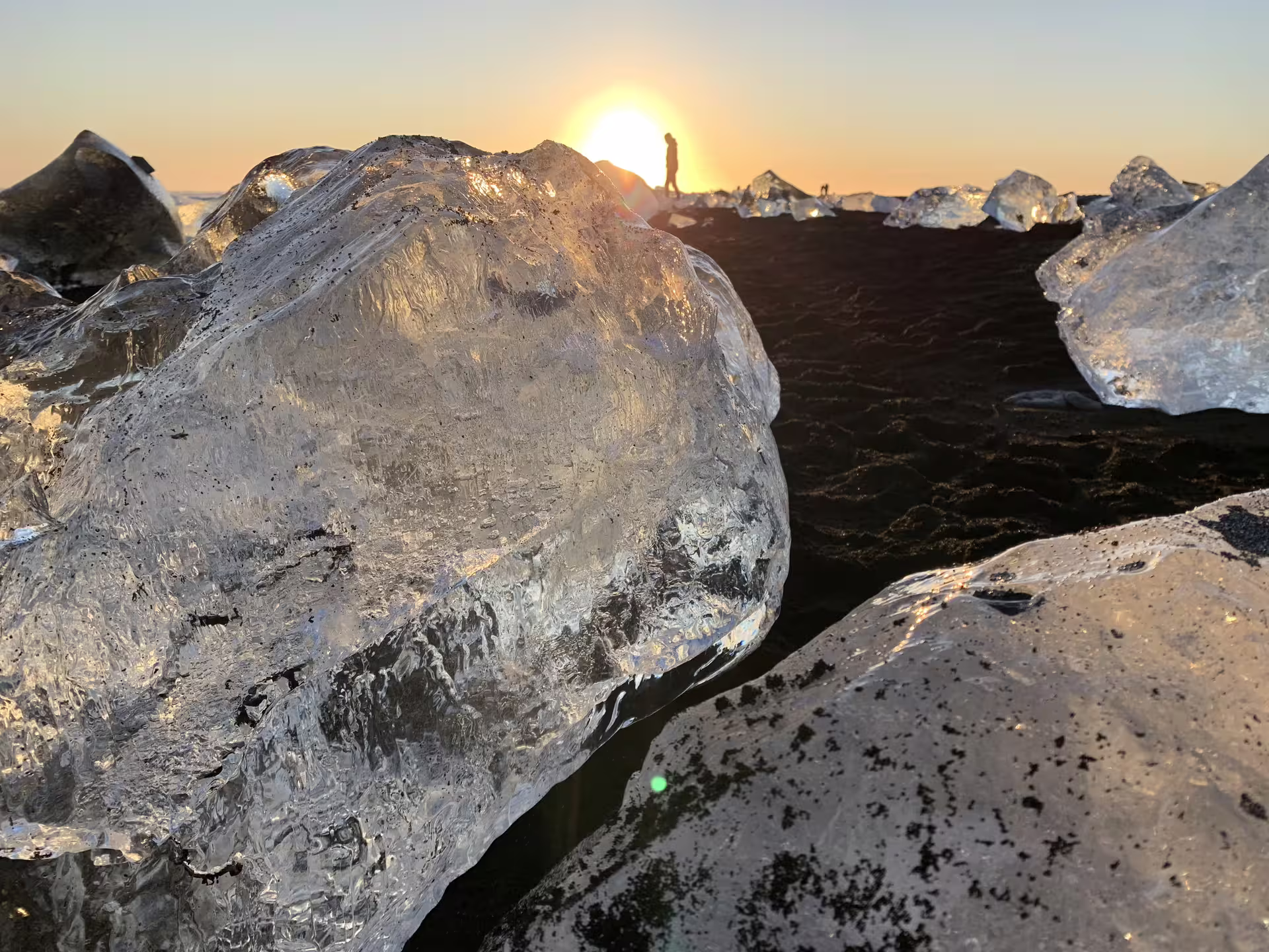 Large glacial ice chunks on black sand beach with lone silhouette against golden sunset in Iceland