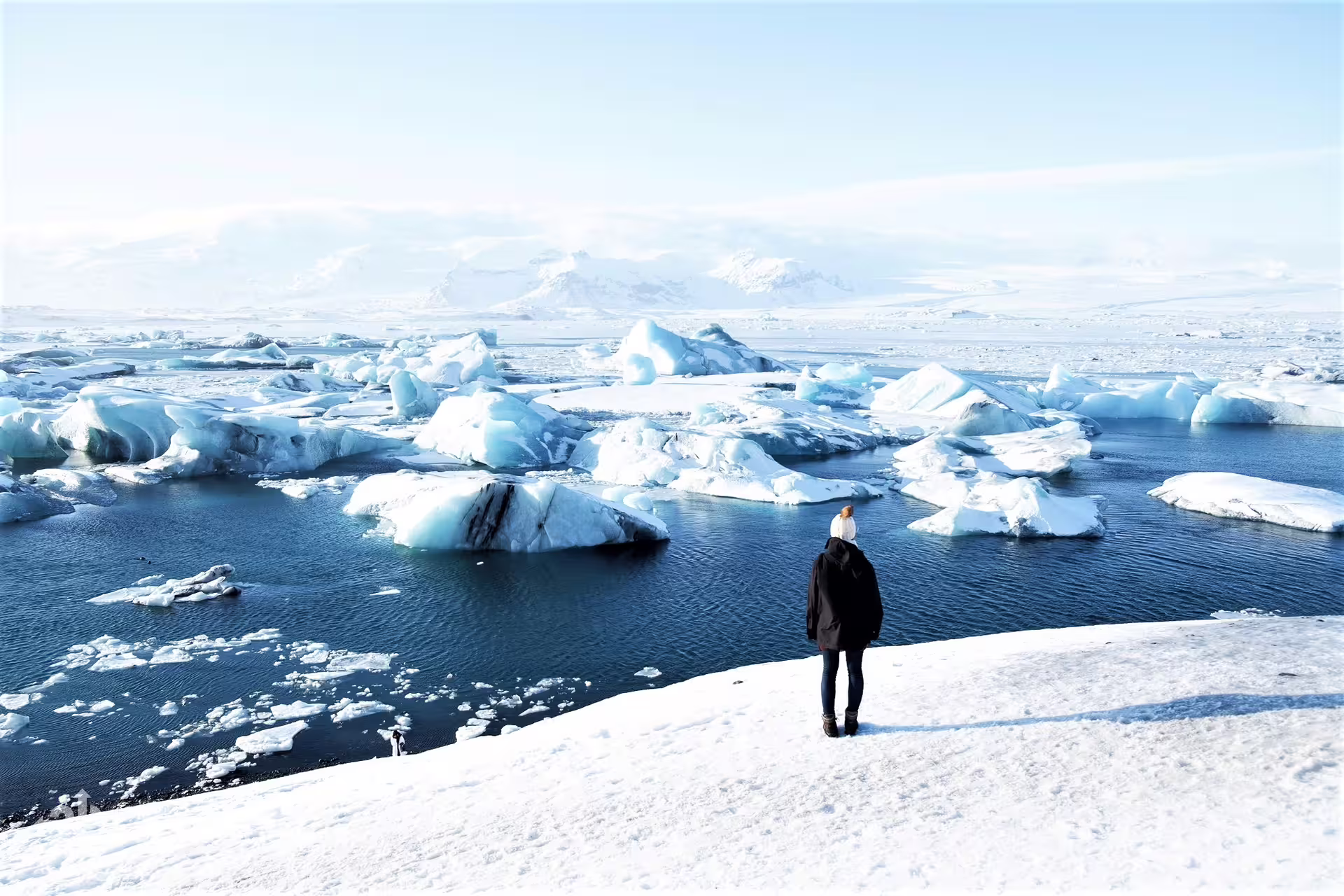 Person standing on snowy shore overlooking Jökulsárlón glacier lagoon with floating icebergs in Iceland