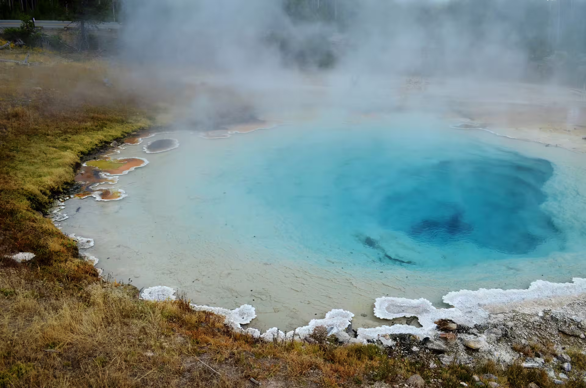 Steaming turquoise hot spring with mineral deposits and colorful microbial mats at edges