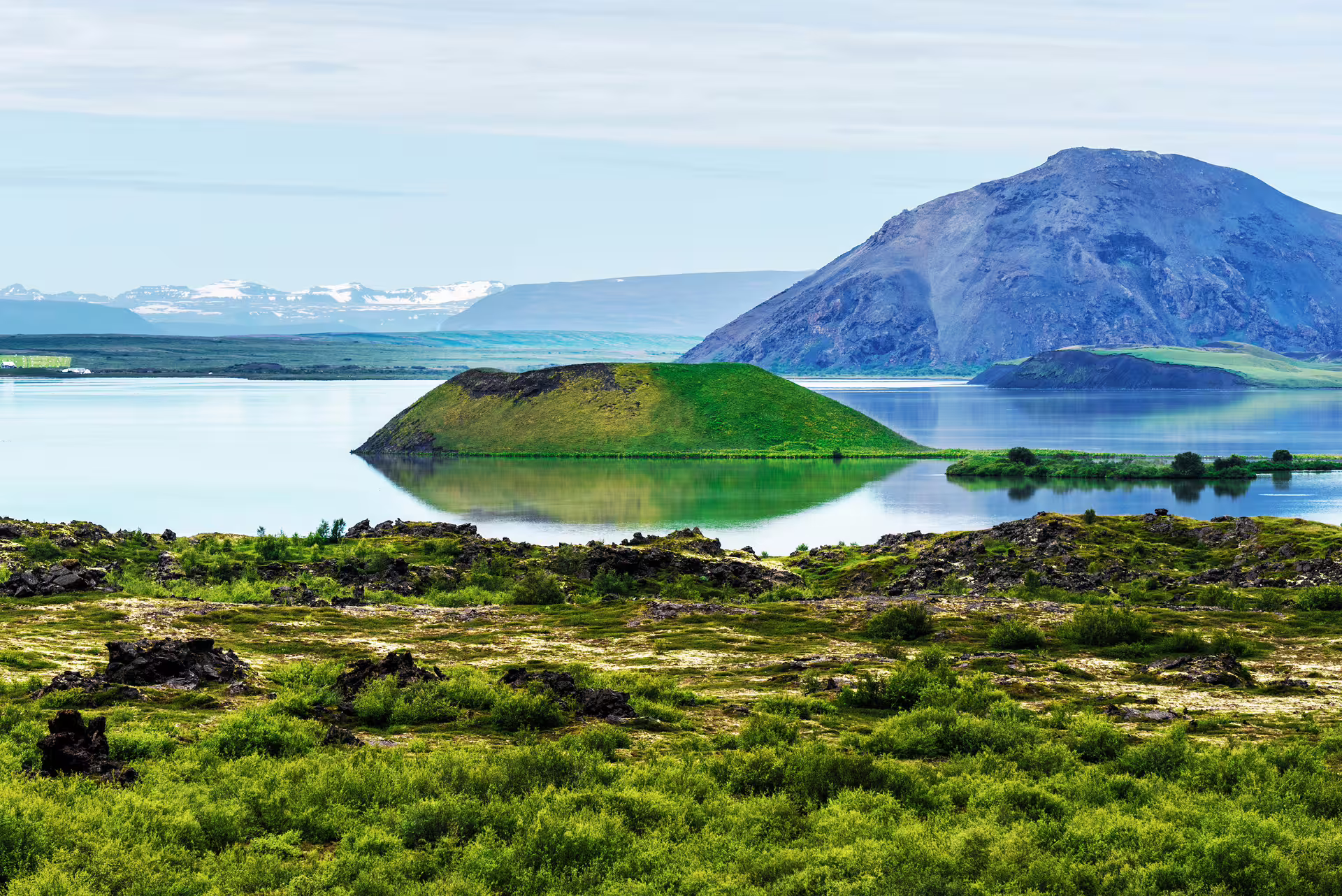 Green volcanic crater island reflected in calm Lake Myvatn with mountain backdrop, Iceland