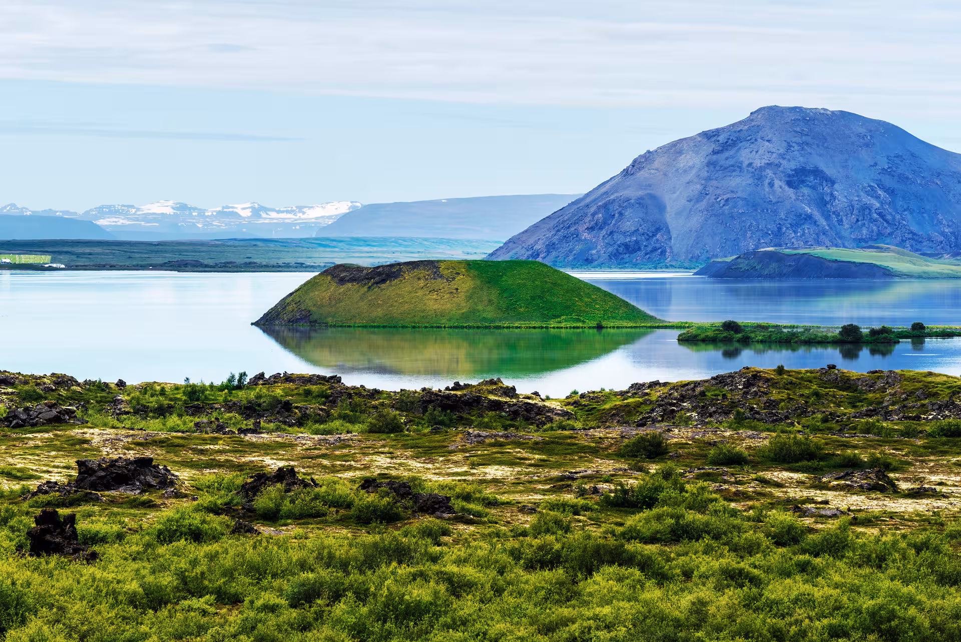 Green volcanic pseudo-crater island reflected in calm Lake Mývatn with mountain backdrop, Iceland