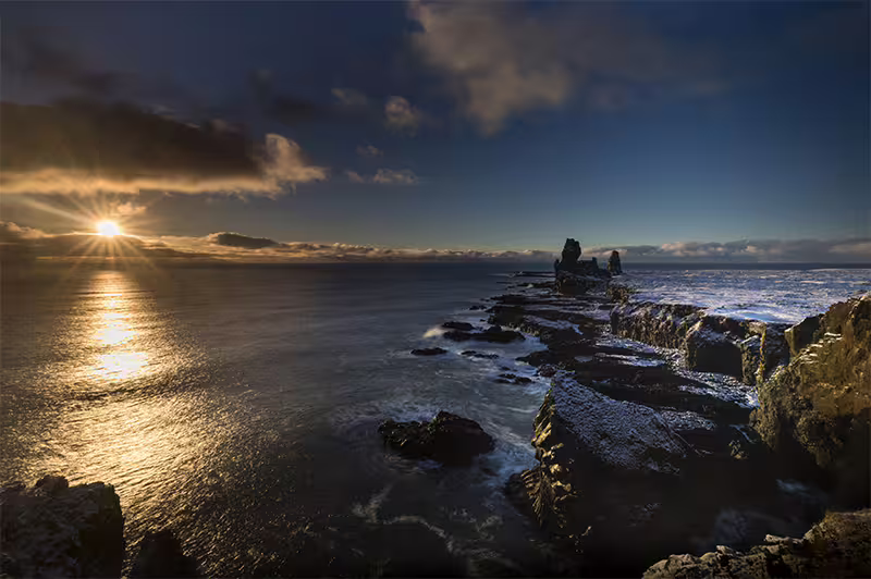 Rocky Iceland coastline at sunset with dramatic cliffs, crashing waves, and golden light reflecting on the ocean