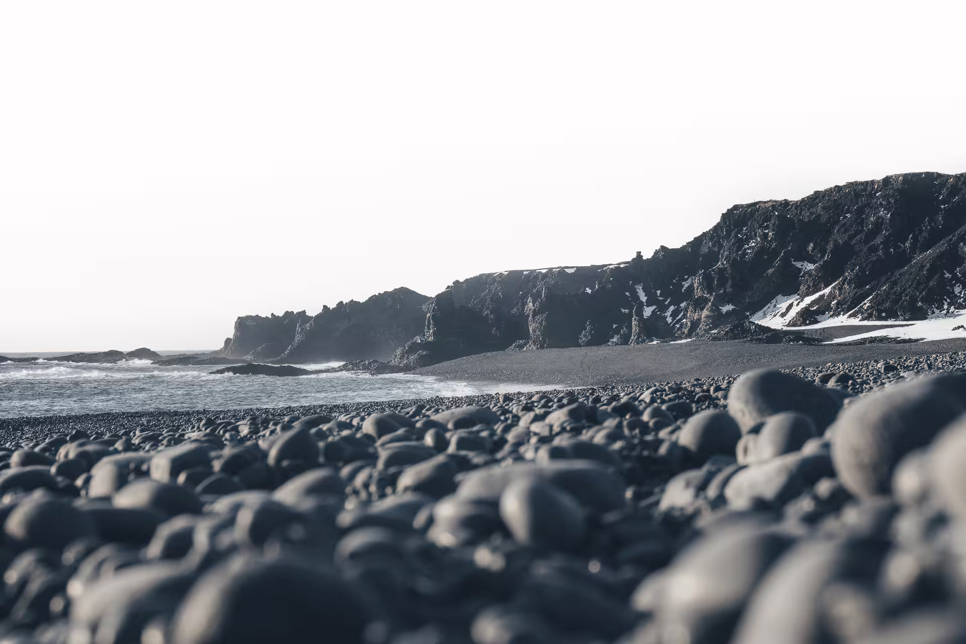 Black pebble beach with dark volcanic cliffs and snow patches along Iceland's rugged coastline