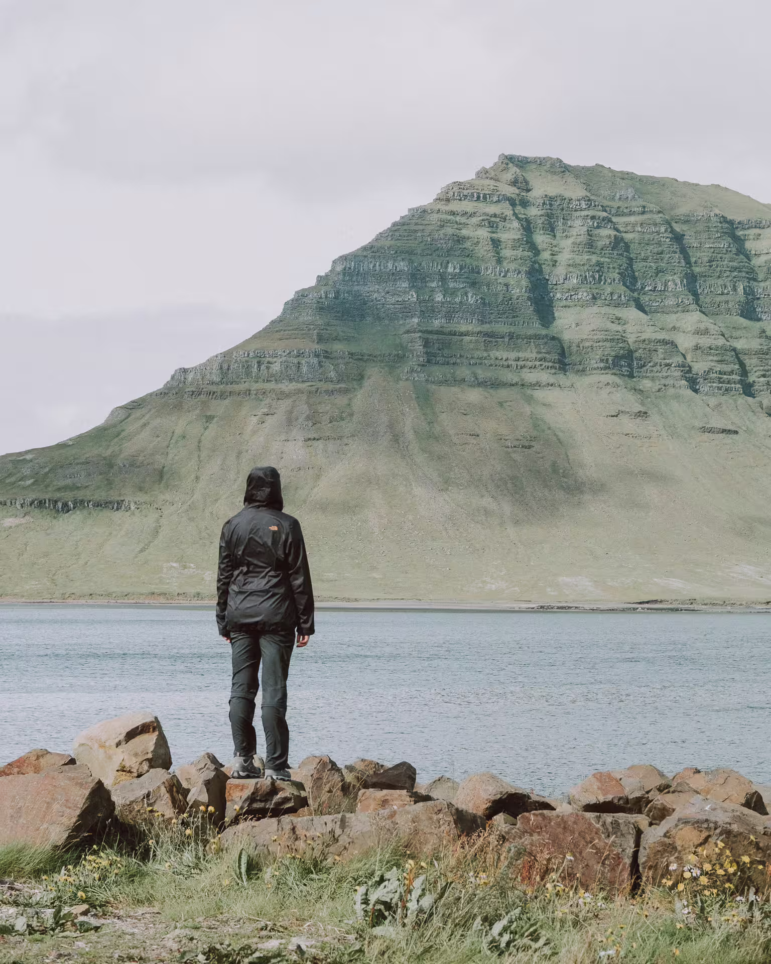 Person in North Face jacket standing on rocks overlooking fjord with layered green mountain in Iceland