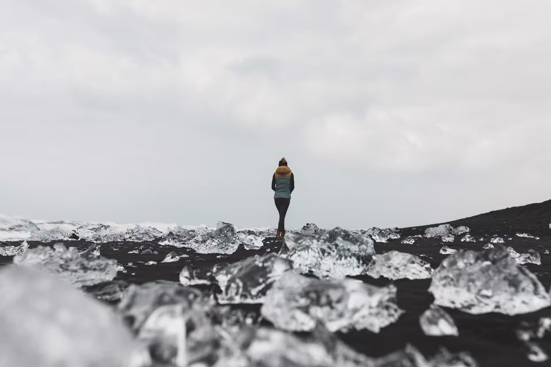 Person standing among glacier ice chunks on Iceland's black sand beach under overcast skies