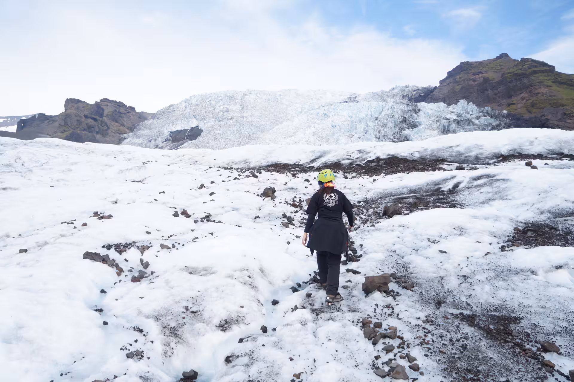 Hiker in black jacket and yellow helmet walking on Iceland glacier with ice formations and mountains