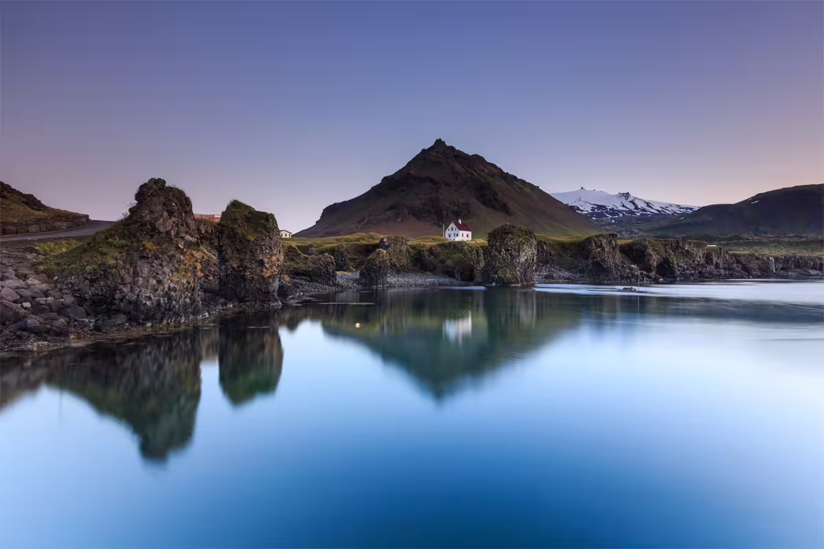 Calm blue lagoon reflecting volcanic rock stacks and a lone white house below a pyramid-shaped mountain in Iceland