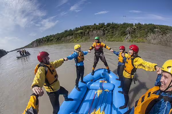Group of rafters in yellow jackets standing on blue raft holding hands on Iceland river