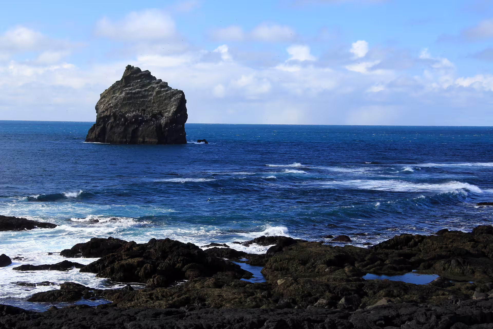 Rocky volcanic coastline with dramatic sea stack rising from blue Atlantic Ocean waters in Iceland