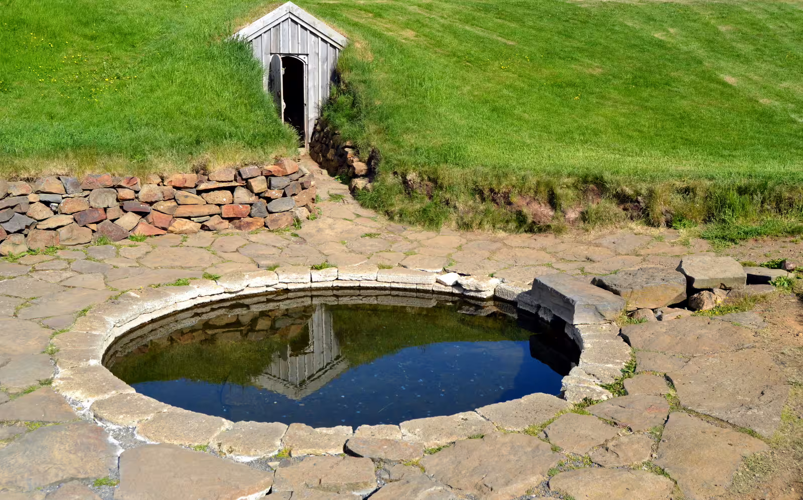 Ancient stone hot spring pool with turf house entrance on green hillside in Iceland
