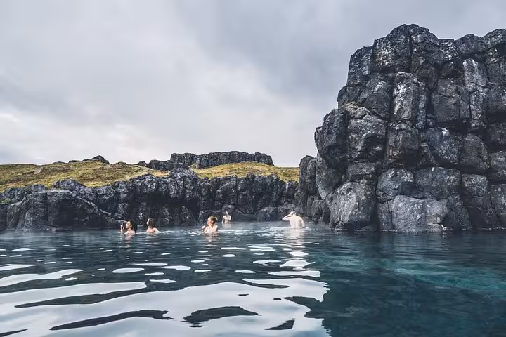 Visitors bathing in a geothermal lagoon surrounded by dark volcanic rocks under cloudy Icelandic skies