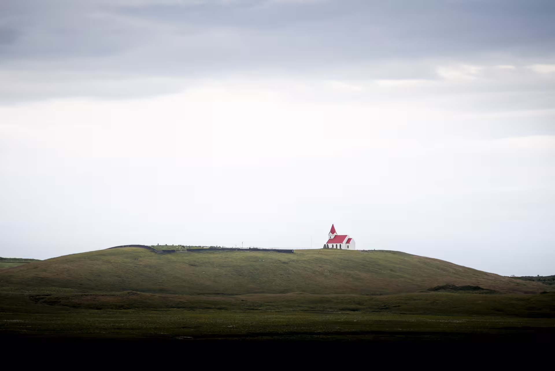 Small white church with red roof on a green hilltop under overcast skies in Iceland