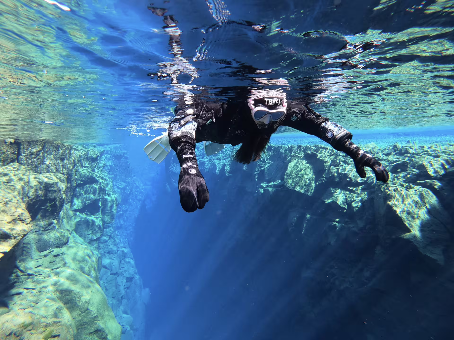 Snorkeler in black drysuit floating above rocky underwater canyon in crystal clear blue water, Iceland