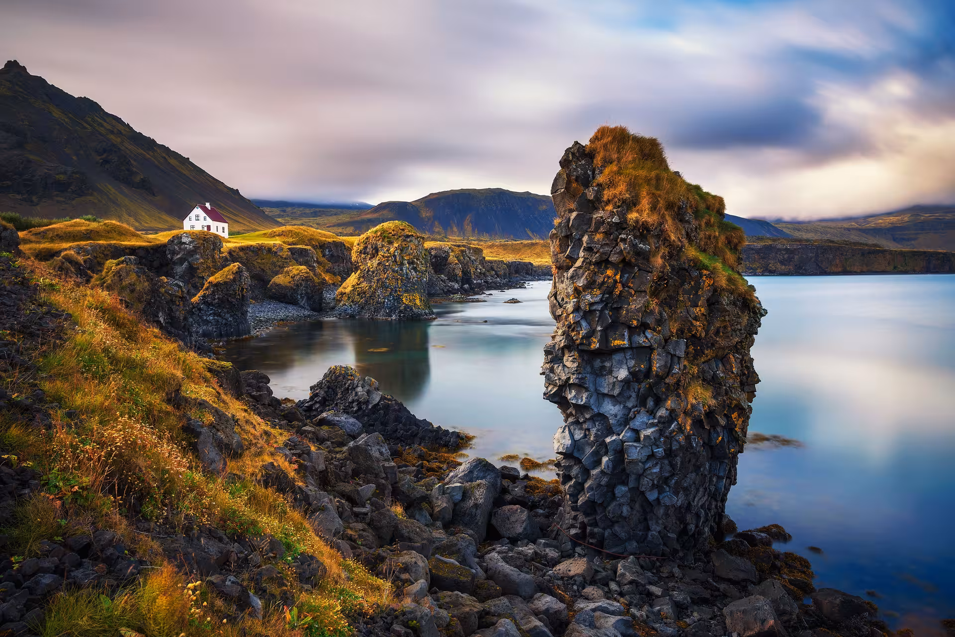 Dramatic Icelandic coastal landscape with volcanic rock formations, calm bay, and lonely white house under moody sky