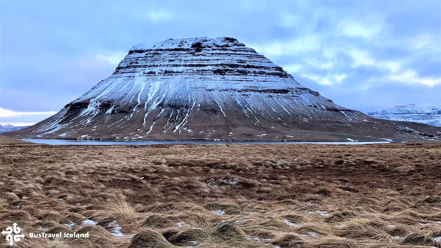 Snow-dusted flat-topped mountain with layered rock face reflected in calm water, Iceland winter landscape