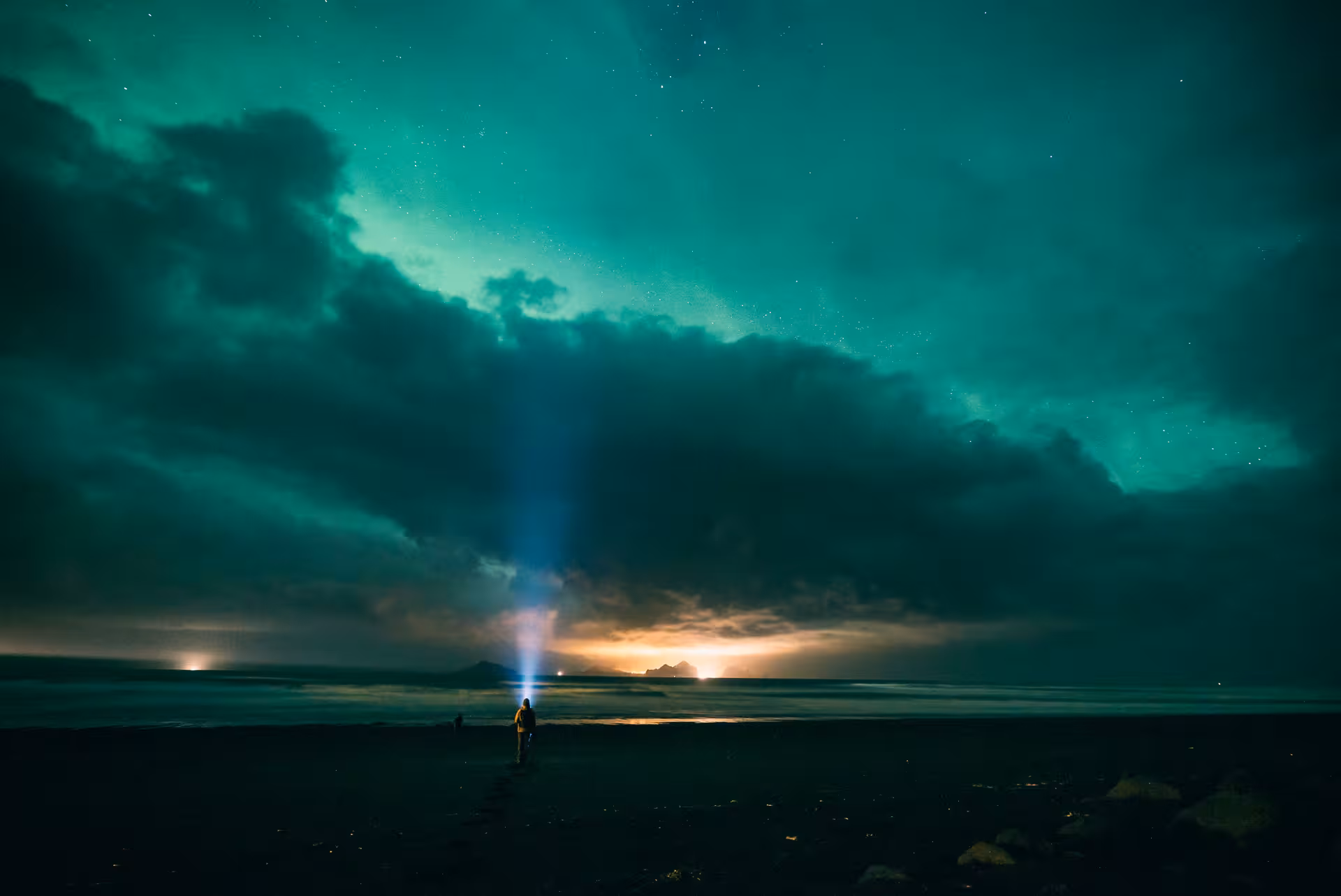 Person with headlamp standing on Iceland's black sand beach under vivid green aurora borealis and dramatic storm clouds