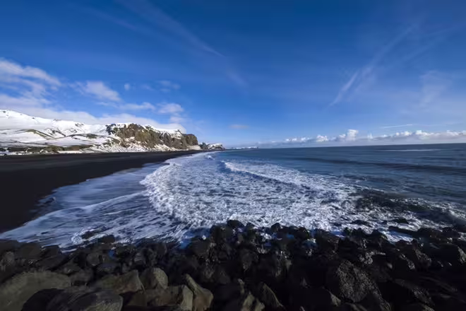 Black sand beach with crashing waves, snow-capped cliffs, and blue sky on Iceland's South Coast