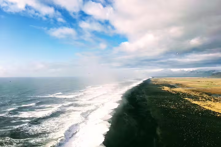 Aerial view of Iceland's black sand beach coastline with crashing waves and green fields under cloudy sky