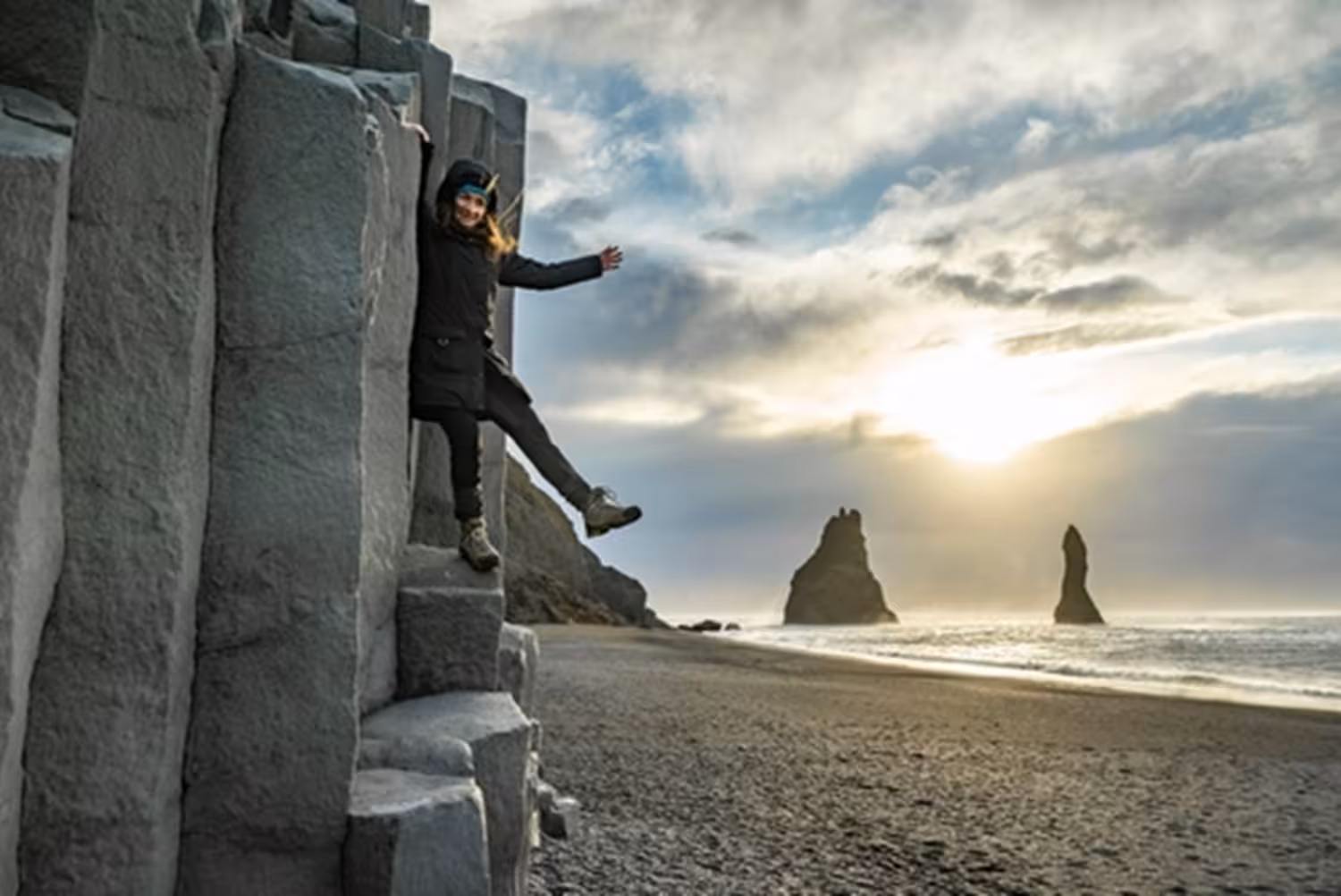 Woman climbing basalt columns on Iceland's black sand beach with sea stacks at sunset