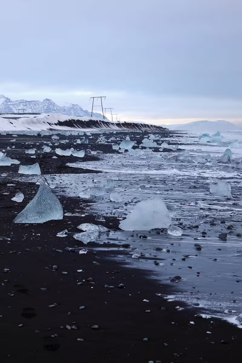 Ice chunks scattered on black sand beach with snowy mountains in background, Iceland South Coast