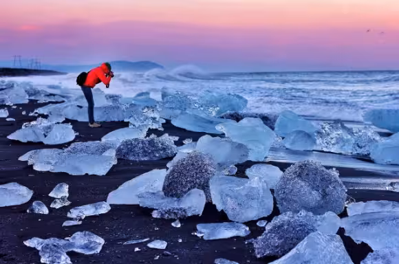 Photographer capturing ice chunks on Iceland's black sand beach at sunset with pink sky