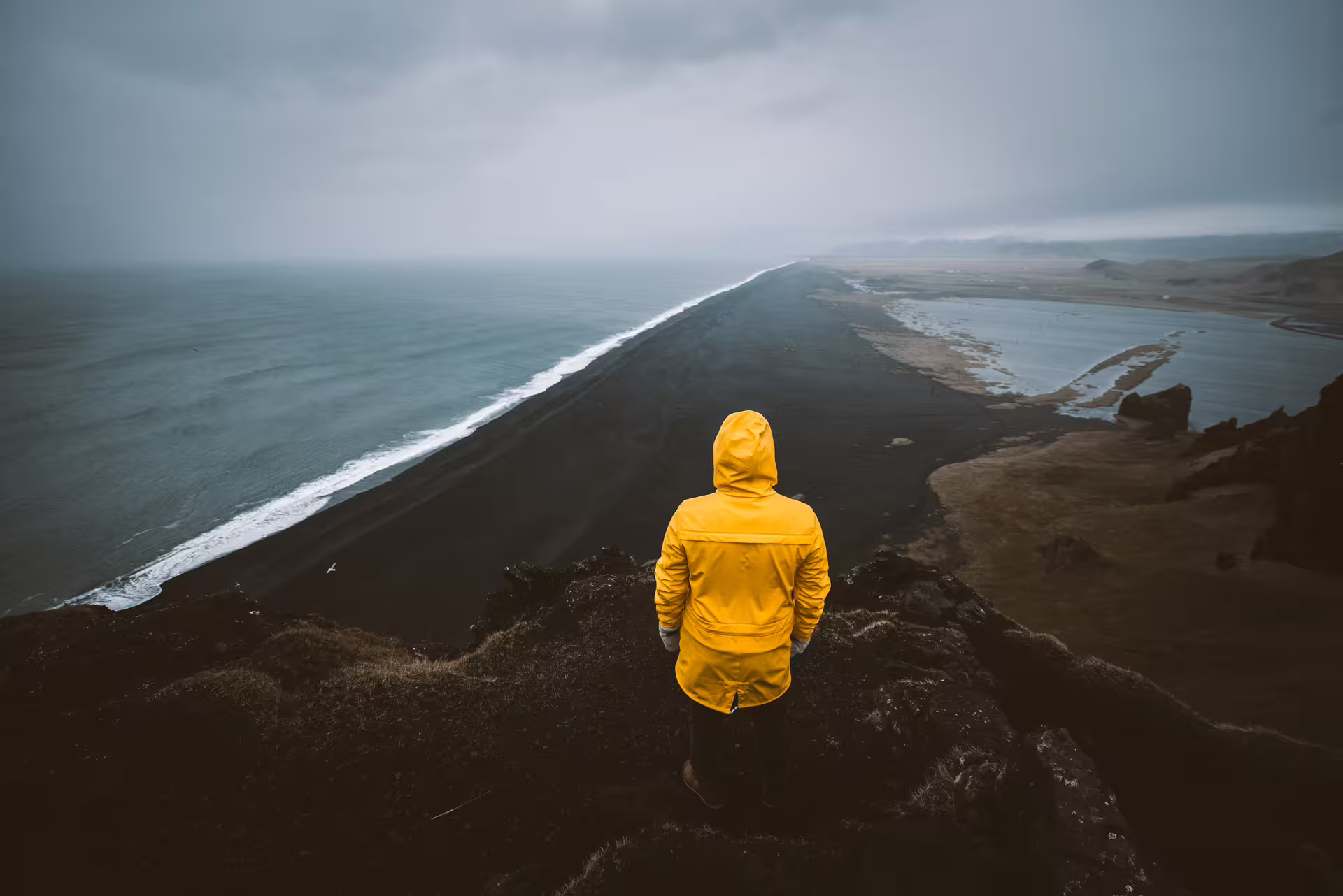 Person in yellow raincoat overlooking Iceland's black sand beach and South Coast from a clifftop