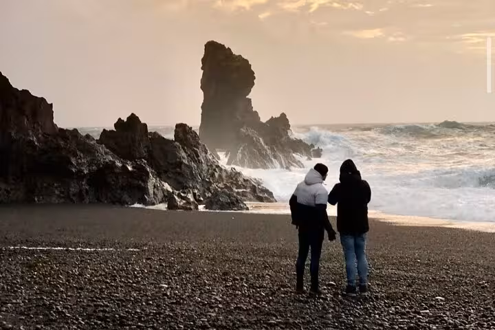 Two visitors standing on Iceland's black sand beach with dramatic sea stack and crashing waves at sunset