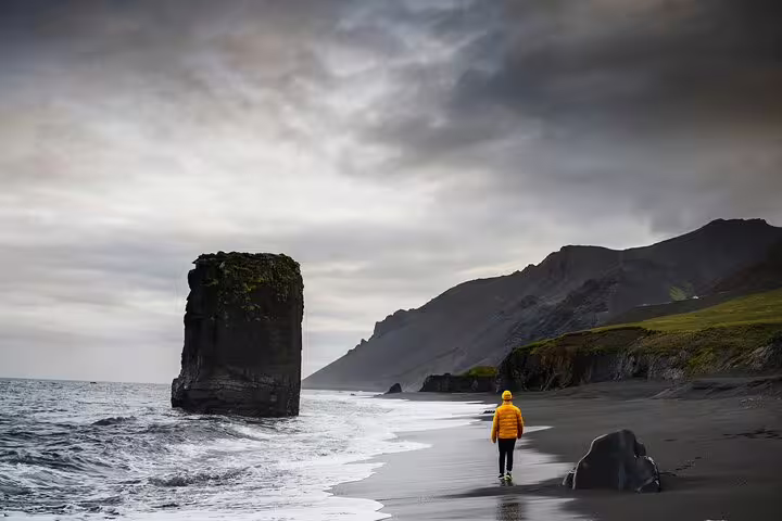 Person in yellow jacket walking on Iceland's black sand beach beside a towering sea stack on the South Coast