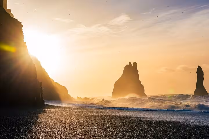 Black sand beach with dramatic basalt sea stacks rising from crashing waves at golden sunset, Iceland South Coast