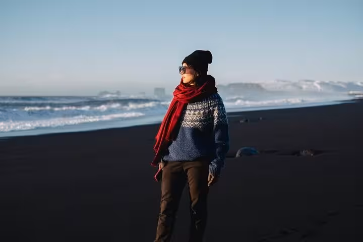 Traveler in winter sweater and red scarf standing on Iceland's black sand beach with ocean waves