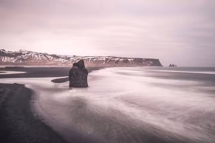 Black sand beach with volcanic rock and silky waves on Iceland's South Coast, snow-capped cliffs in background