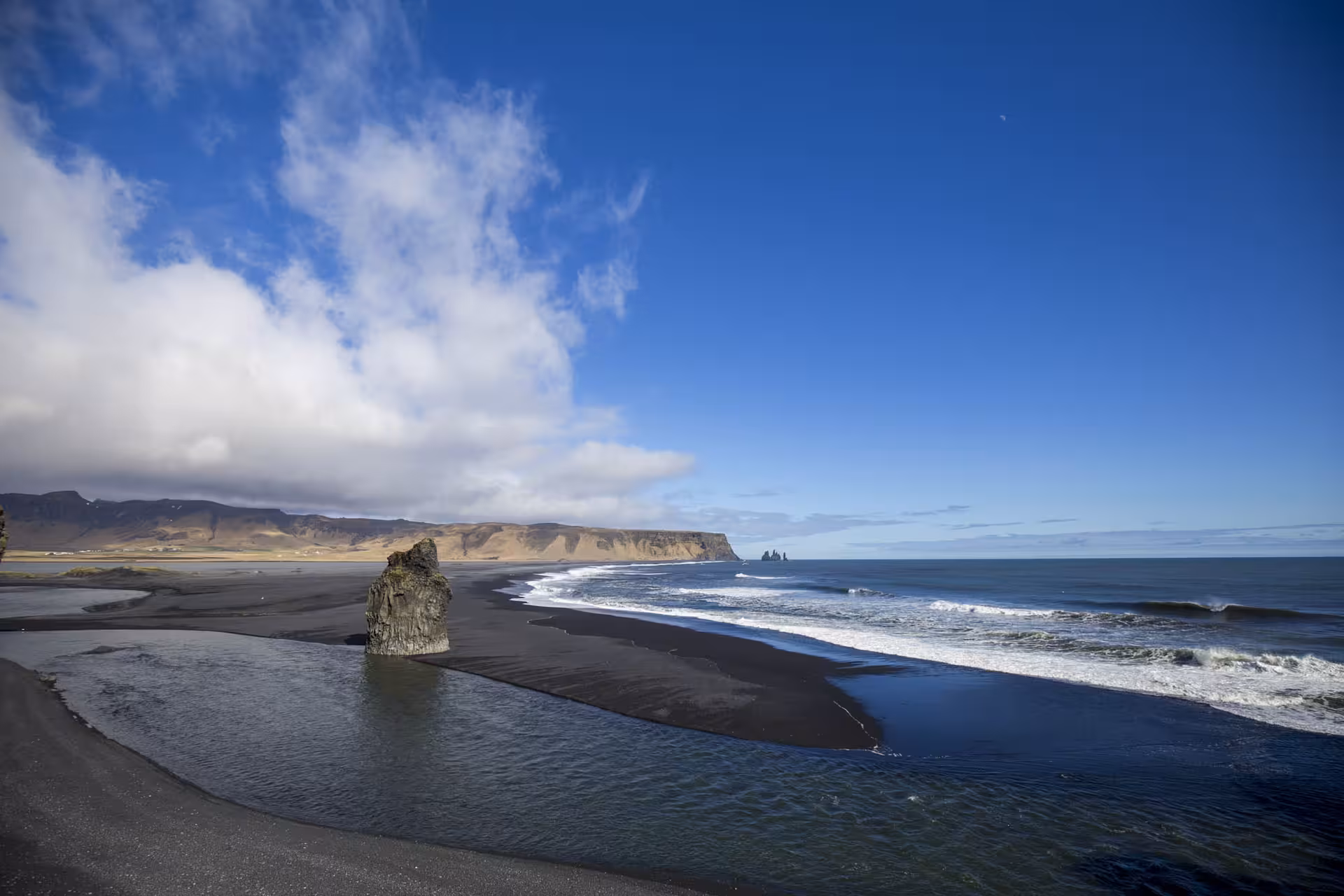 Black sand beach with volcanic rock stack, ocean waves, and distant sea stacks on Iceland's south coast