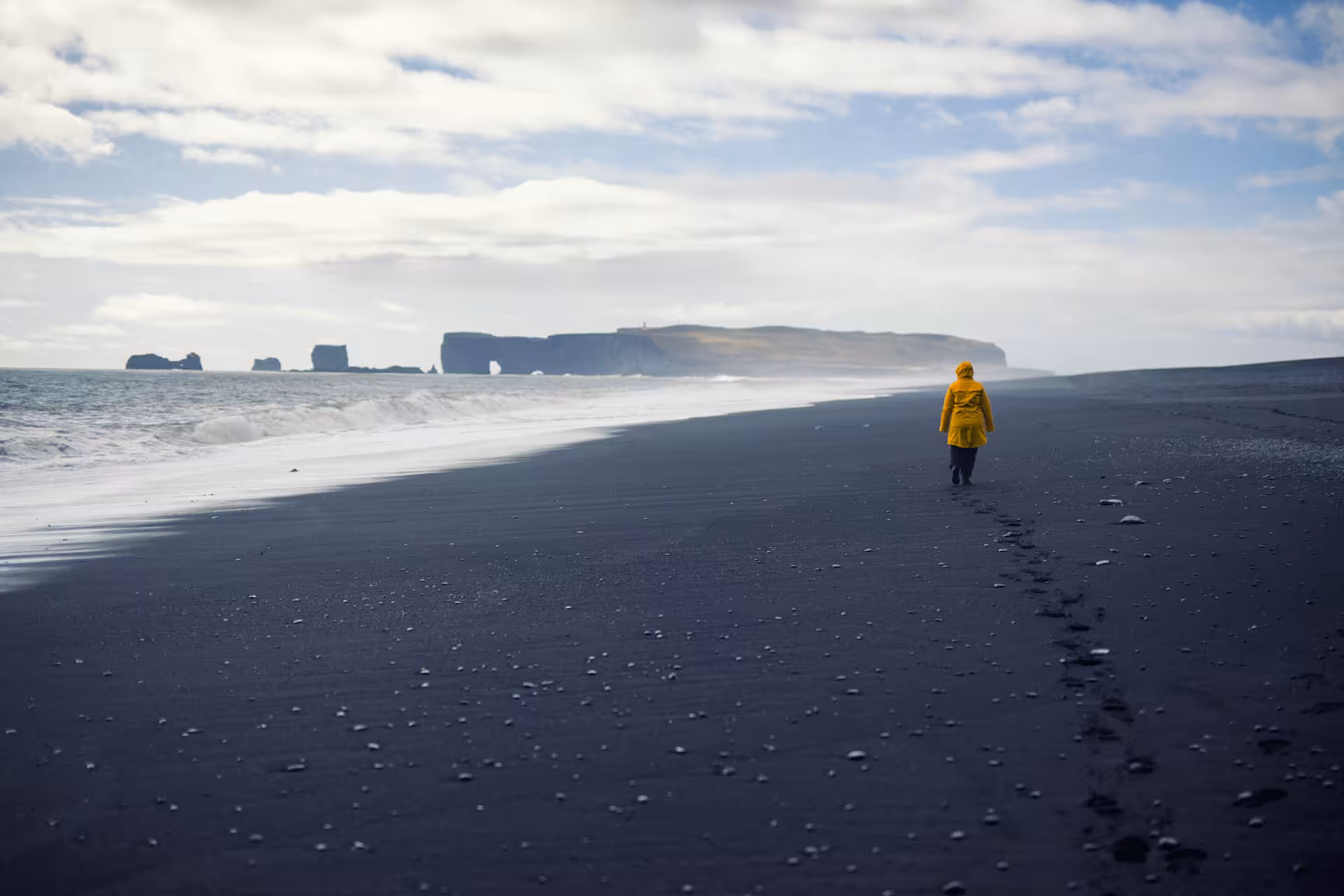 Person in yellow jacket walking on Iceland's black sand beach with dramatic sea stacks in background
